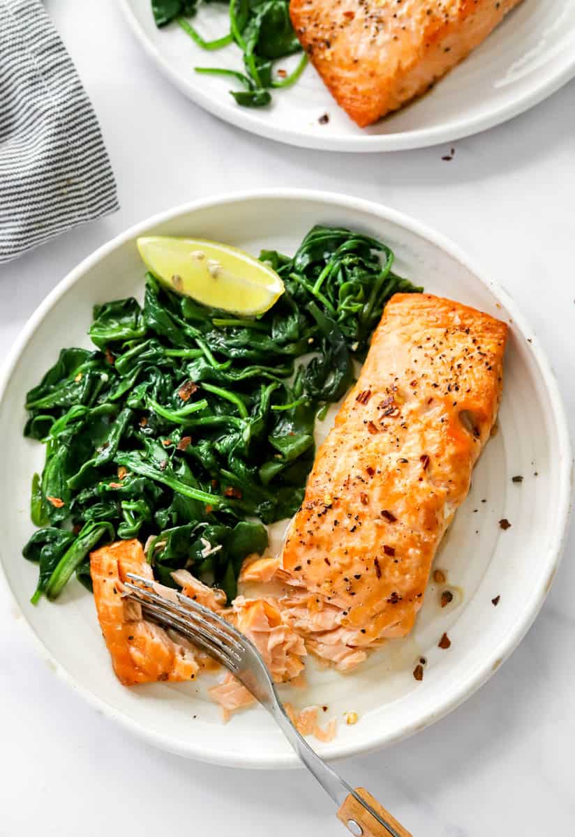 Air fryer salmon on a round white plate with cooked greens with a fork cutting into the salmon.