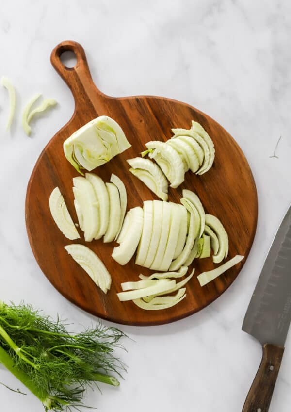 Round wooden cutting board with sliced raw fennel on it with a knife and a fennel build in front of it.