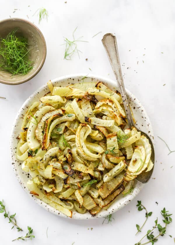 Round white plate of cooked fennel with a spoon on the plate and a bowl of fennel pronged behind it.