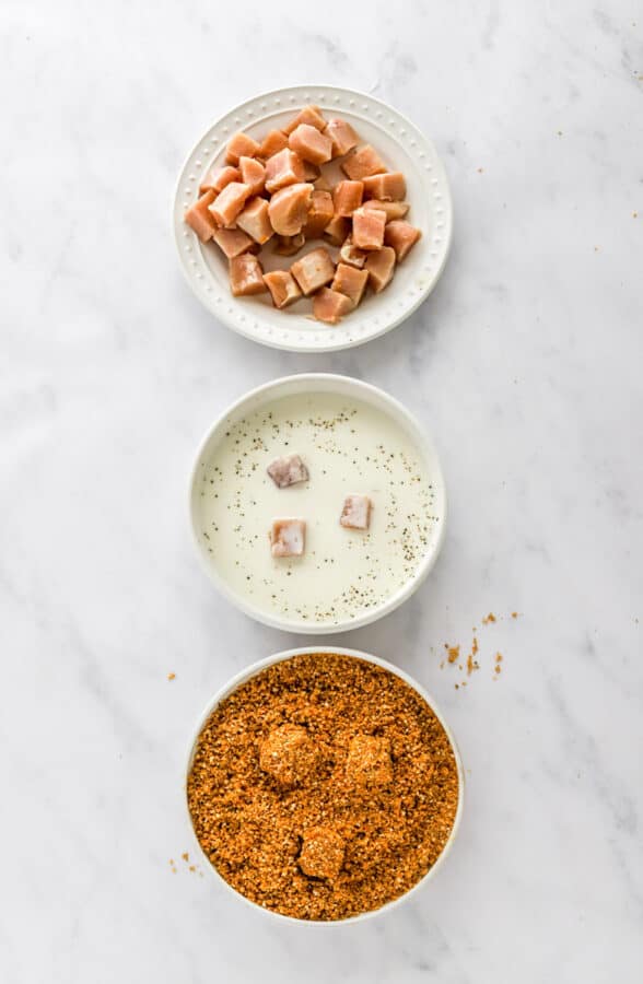 Plate of raw, cubed chicken with a bowl of seasoned milk and a bowl of breadcrumbs in front of it.