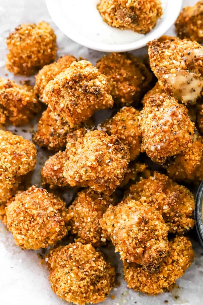 A bunch of golden cooked air fryer popcorn chicken in a pile on a plate with a bowl of white sauce behind it.