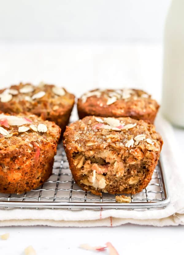 4 muffins with oats and sugar on top of them on a silver wire rack with a jug of milk behind them.