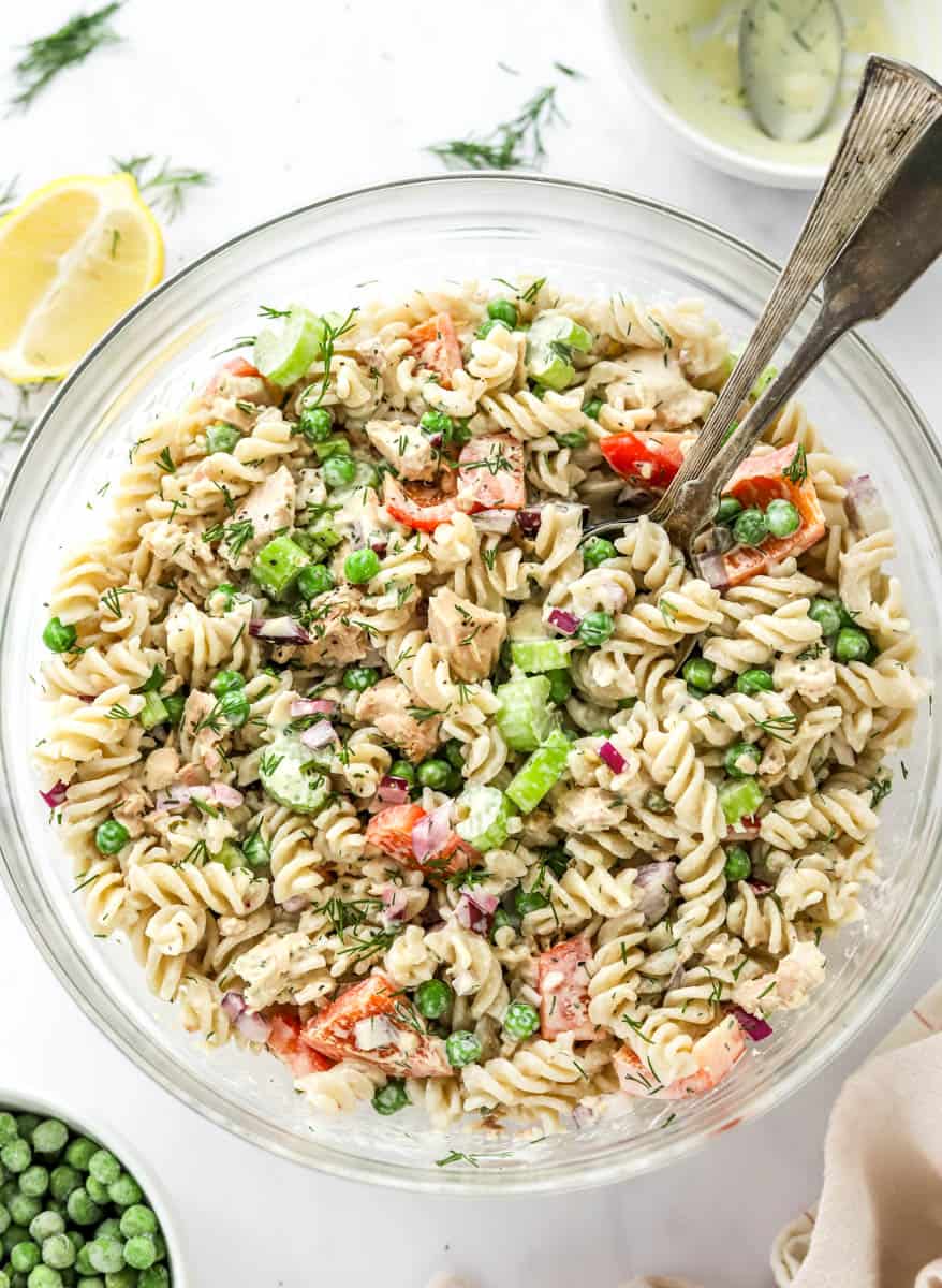 Healthy tuna pasta salad in a round glass bowl with silver serving spoons in it with a cut lemon and more creamy sauce behind it. 