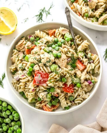 Tuna pasta salad in a bowl with another bowl of it behind it and a bowl of peas in front of it.