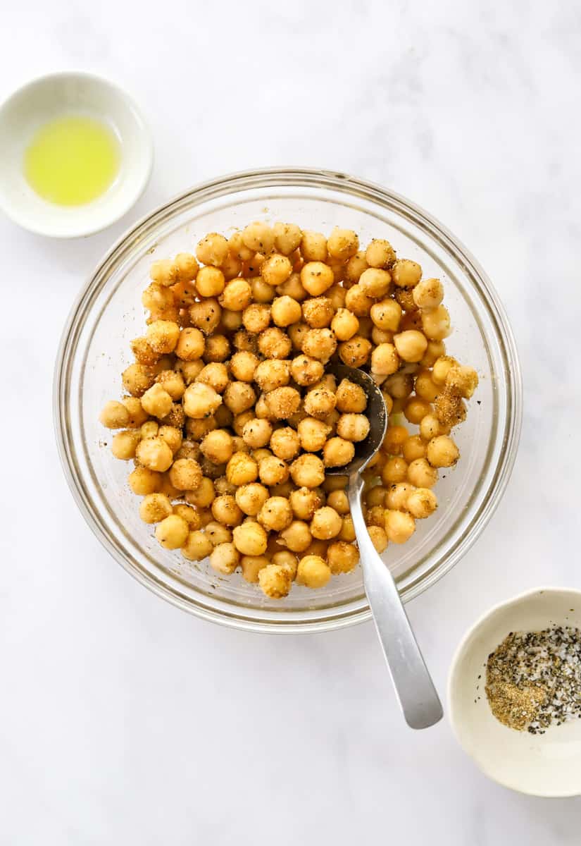 Round glass bowl with chickpeas seasoned with spices in it with a small bowl of oil behind it and a bowl of spices in front of it.