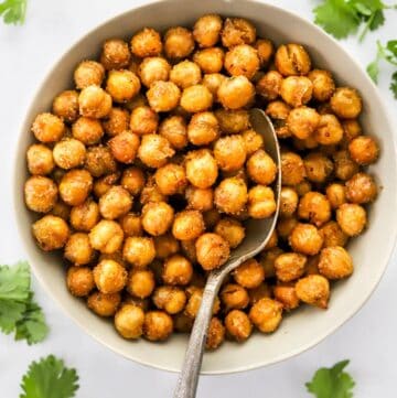 Light brown bowl with air fryer chickpeas in it with a spoon in the bowl and torn green herbs scattered around the bowl.