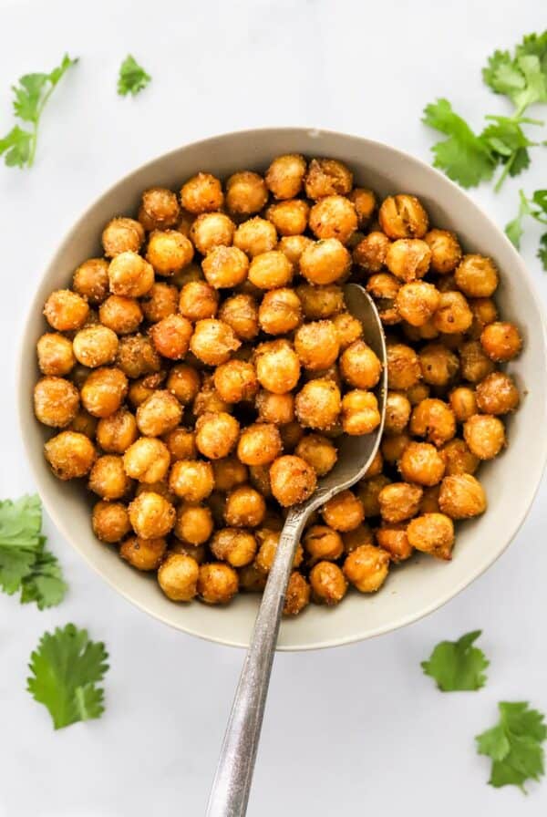 Light brown bowl with air fryer chickpeas in it with a spoon in the bowl and torn green herbs scattered around the bowl.