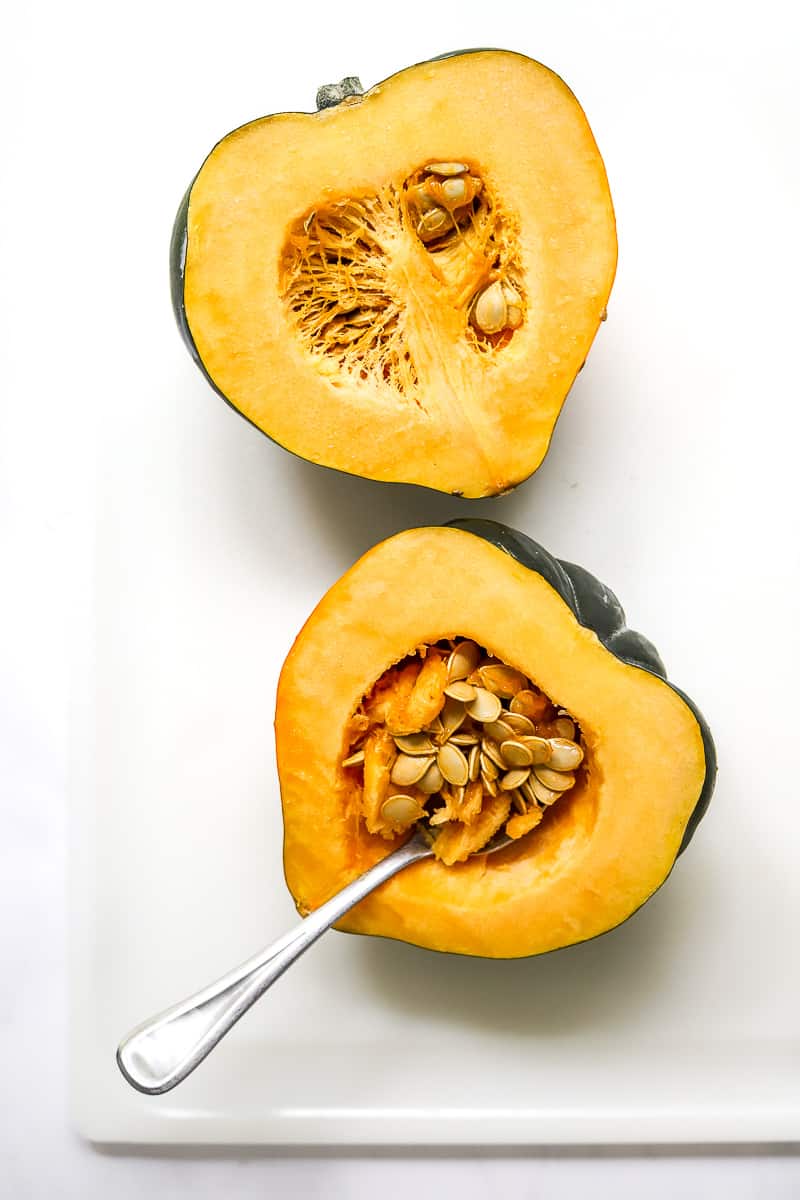 Cut acorn squash halves on a white board with a spoon scooping seeds out of one of the squash.