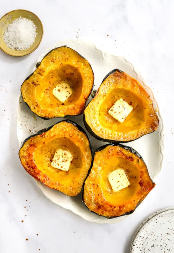 Oval platter with roasted acorn squash on it with pats of butter on the squash with a yellow dish of salt behind it.