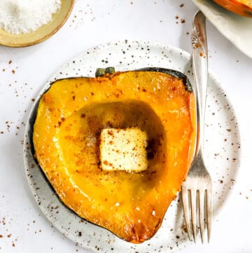 Round specked plate with a half of squash with butter on it with another plate of squash and small yellow bowl of salt behind it.