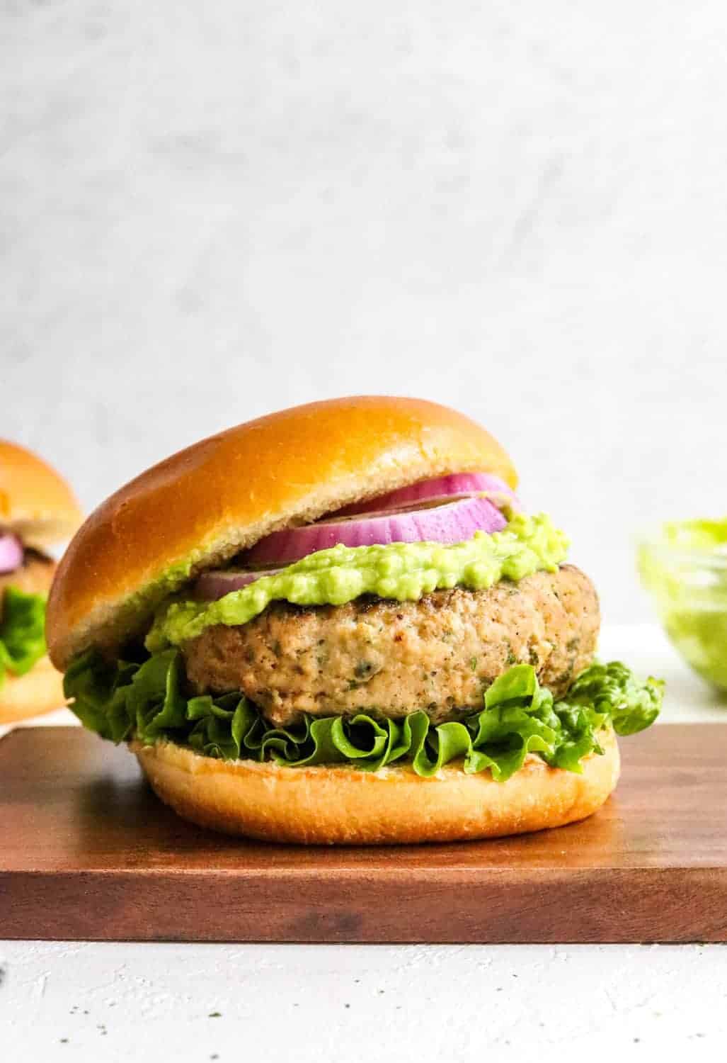 Healthy gluten free turkey burger in between a burger bun with purple onion and mashed avocado on it with another burger behind it. 