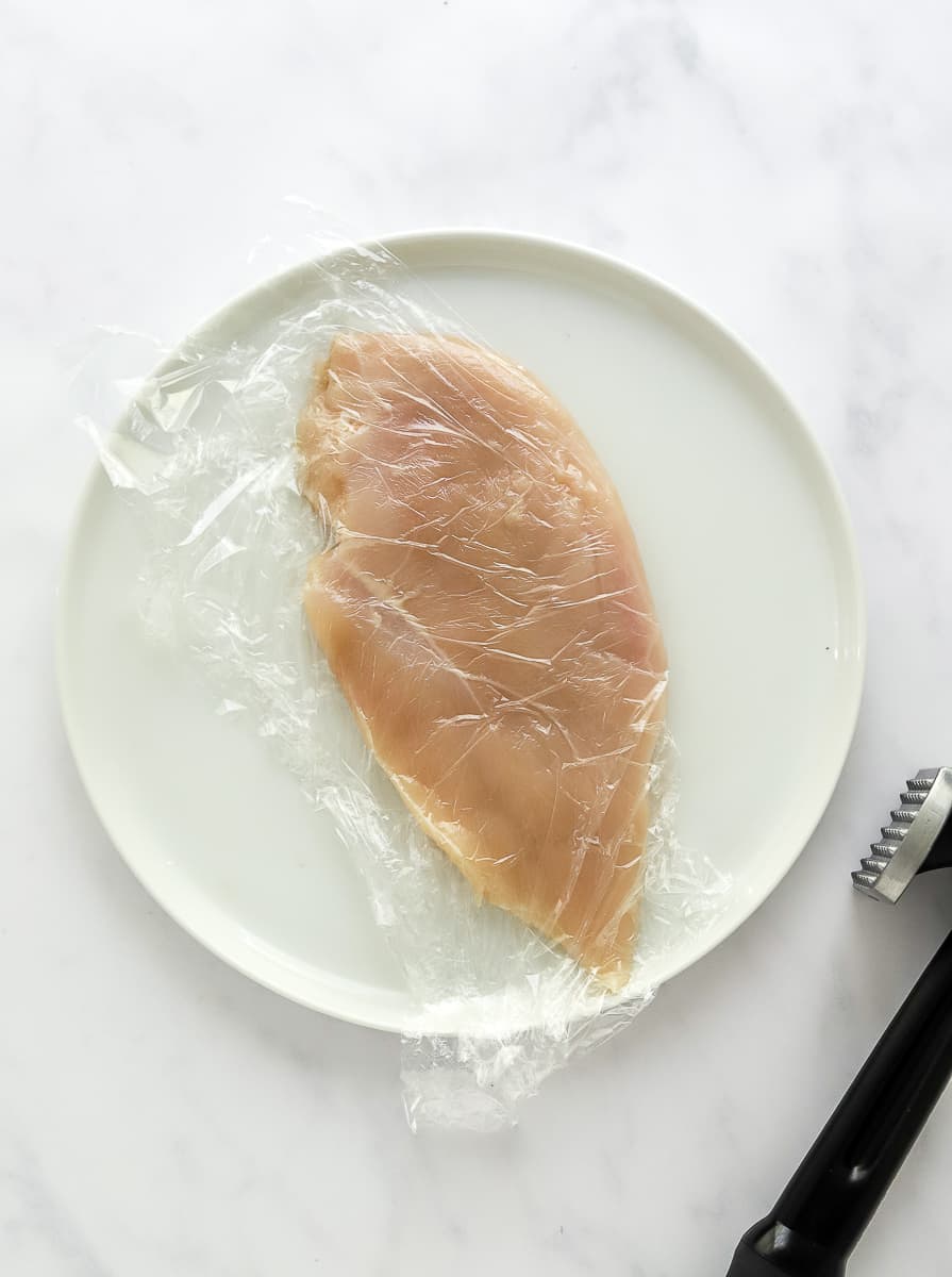Raw piece of chicken in plastic wrap on a white plate with a meat mallet next to it.