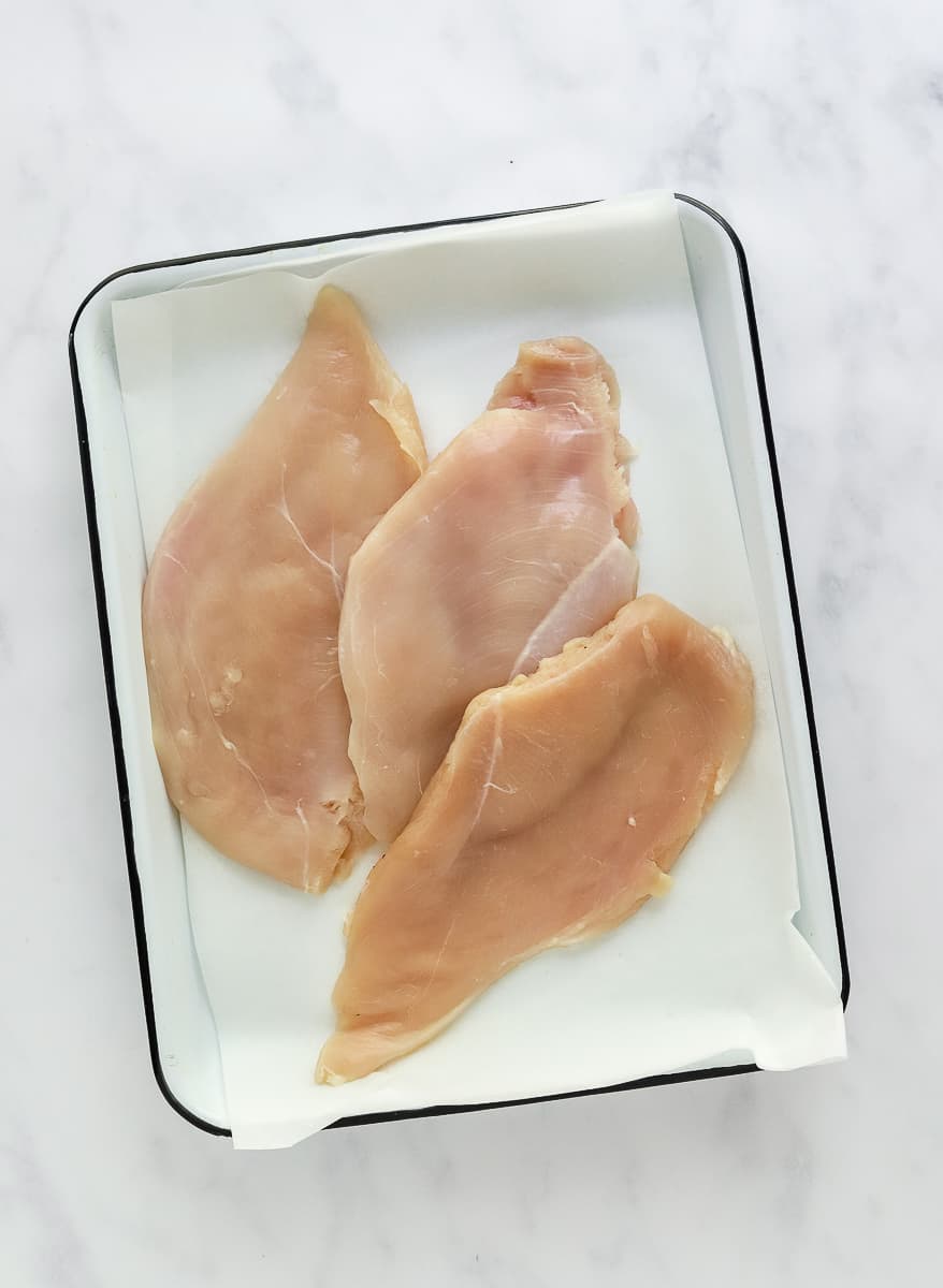 3 uncooked chicken cutlets on a white baking sheet.
