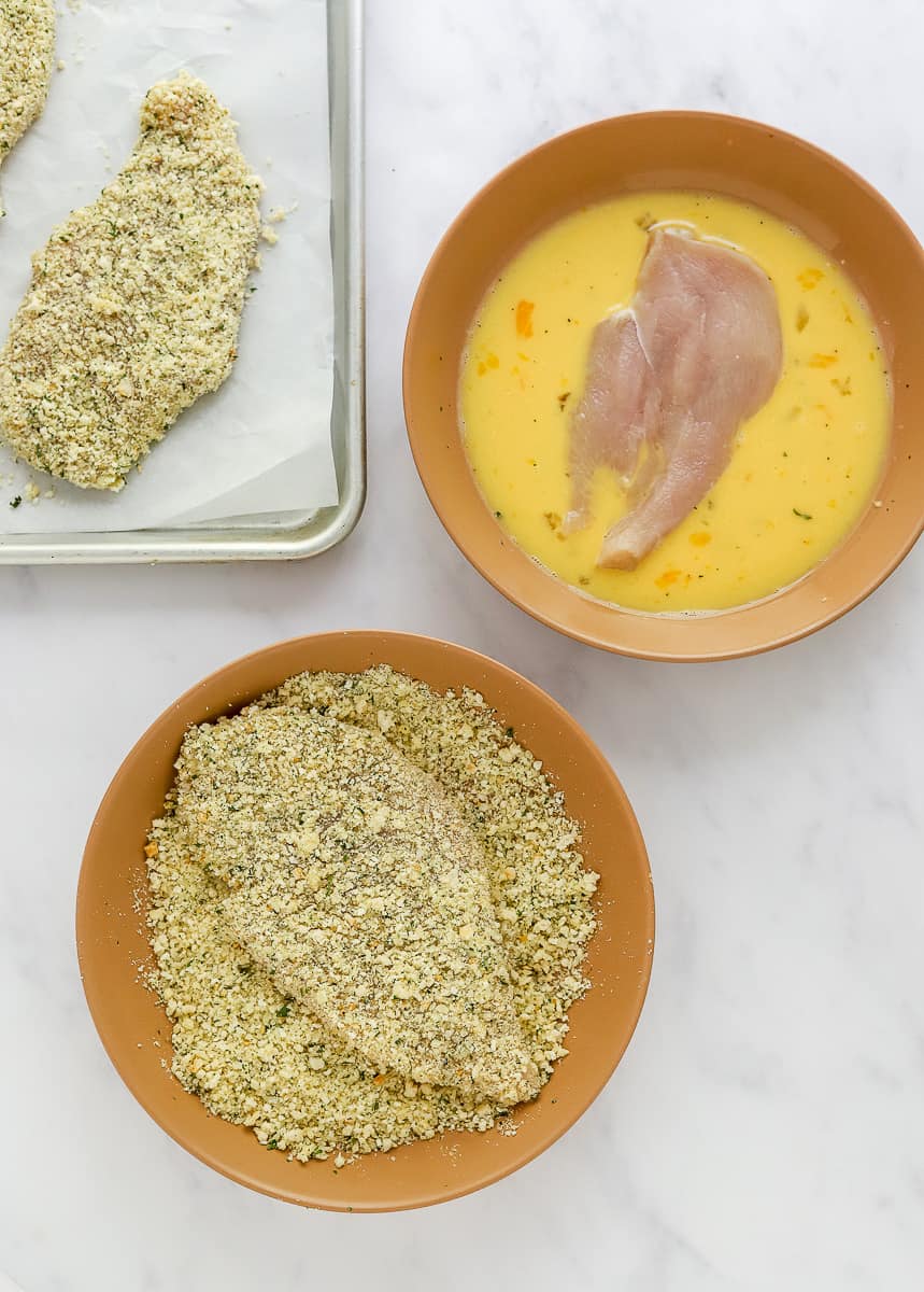 2 brown bowls with on filled with whisked eggs with a piece of raw chicken in it with the other bowl filled with breadcrumbs and a pieces of crusted chicken in with with more breaded chicken behind it.