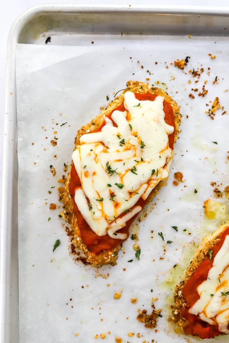 Golden, crusted chicken parmesan with red sauce and melted cheese on top of it with another piece of chicken Parmesan in front of it on white paper on a baking sheet.