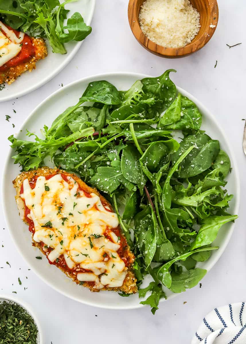 Round white plate fill with greens and a piece of chicken Parmesan with another plate of it and a wooden bowl of shredded cheese behind it.
