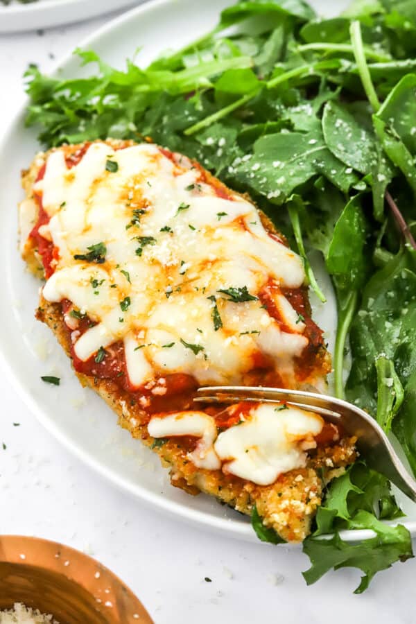 Chicken parmesan on a plate with a fork cutting into it with mixed greens next to it on the plate.