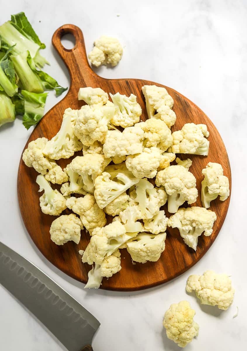 Round wooden cutting board with chopped cauliflower on it with a kitchen knife next to it and cauliflower leaves behind it.