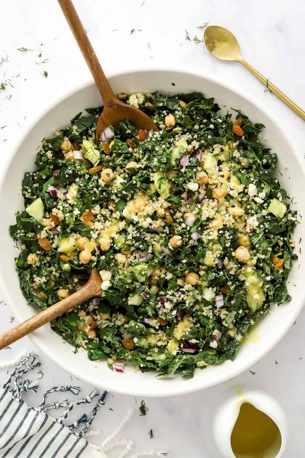 Chopped kale in a large bowl with quinoa chopped veggies and lemon dressing with wooden serving spoons in the bowl with some dressing in a pitcher and a white striped towel in front of it.