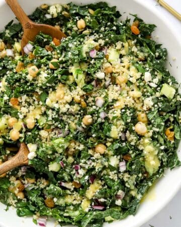 Bowl of chopped kale with quinoa, chickpeas and chopped veggies on it with dressing pour over it with wooden serving spoons in it.