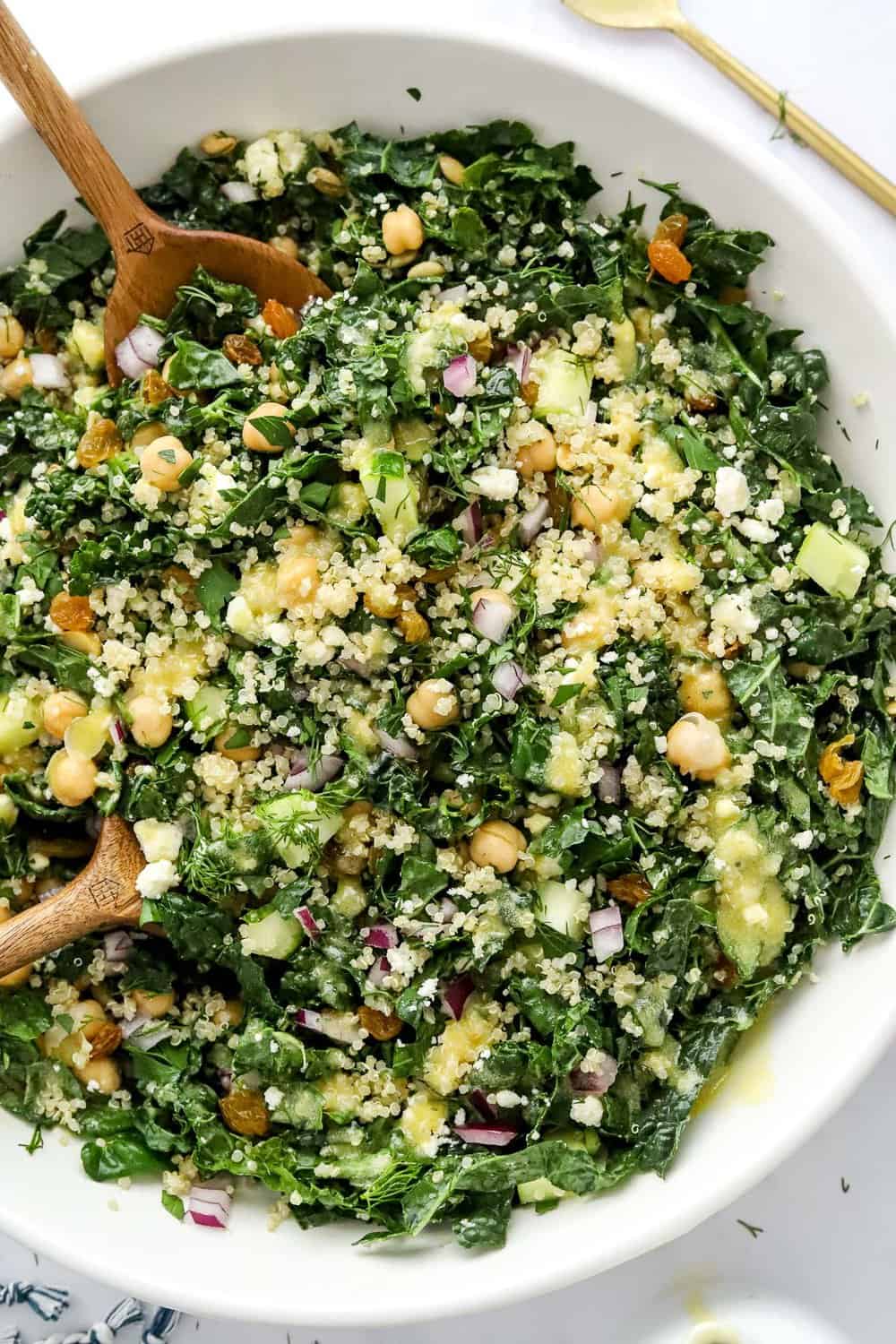 Bowl of chopped kale with quinoa, chickpeas and chopped veggies on it with dressing pour over it with wooden serving spoons in it.