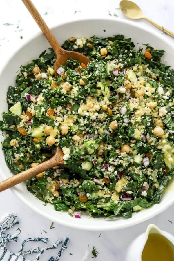 Bowl of sliced green kale topped with veggies, chickpeas and quinoa with spoons in the bowl and a striped towel and more dressing in a pitcher in front of it.