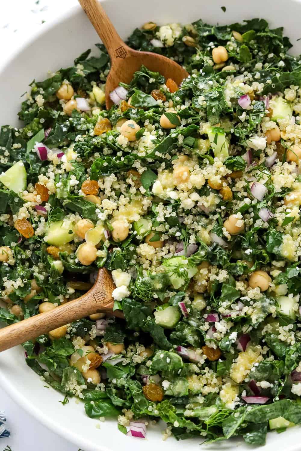 Sliced kale, chickpeas red onion, cucumber and lemon dressing in a serving bowl with wooden salad spoons in the bowl.