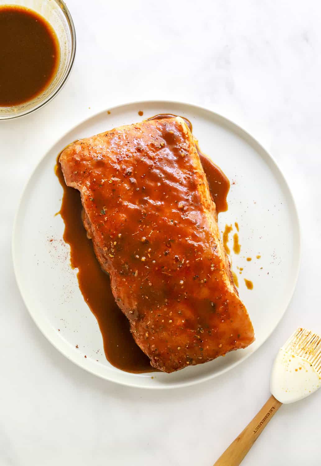 Raw pork loin covered in glaze on a white plate with more glaze in a bowl behind it and a rubber brush in front of it.