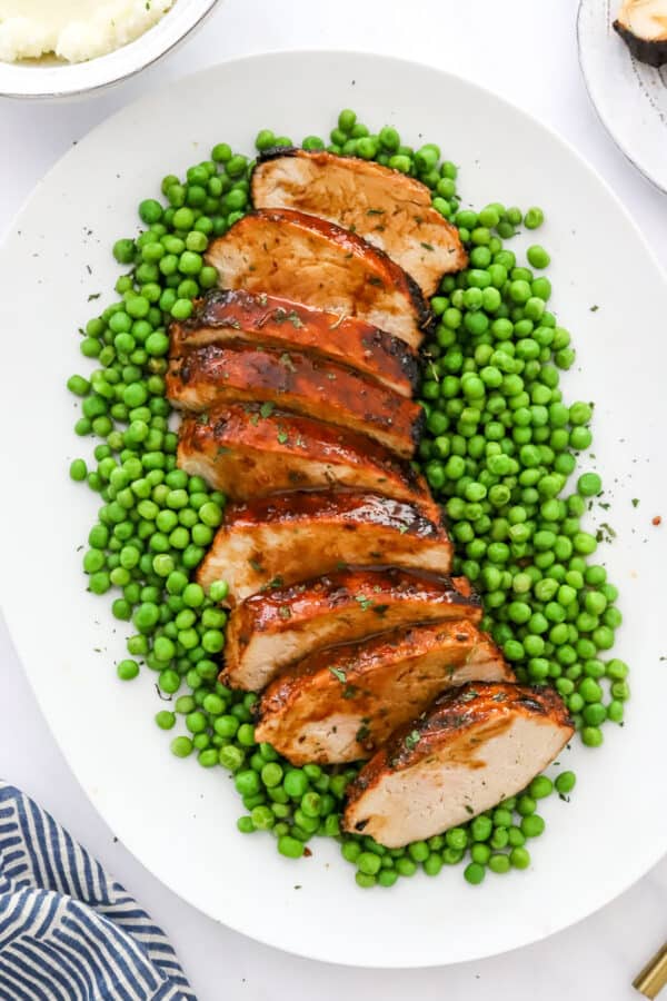 White platter with green peas on it with a sliced pork loin ontop of the peas with a blue linen in front of it and a bowl of mashed potatoes behind it.