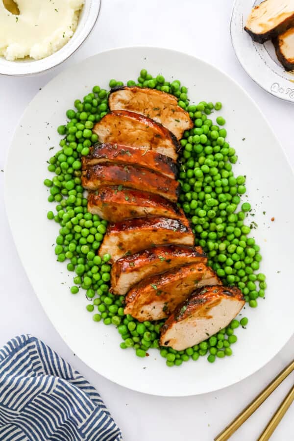 Sliced meat covered in brown glaze on a platter with peas around it with more meat on a plate and a bowl of potatoes behind it.