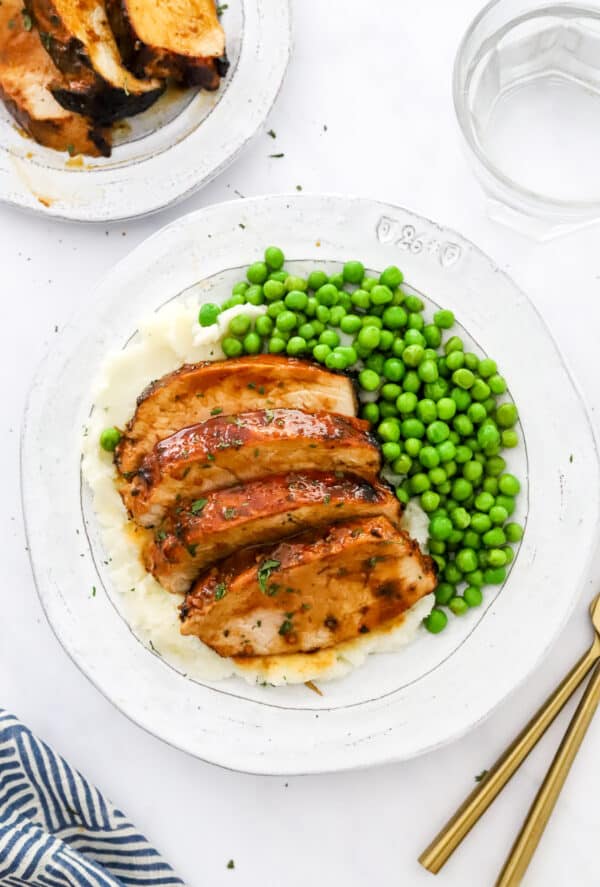 Round plate with sliced brown meat on it on top of mashed potatoes with peas on the plate with another plate of meat and glass of water behind it.
