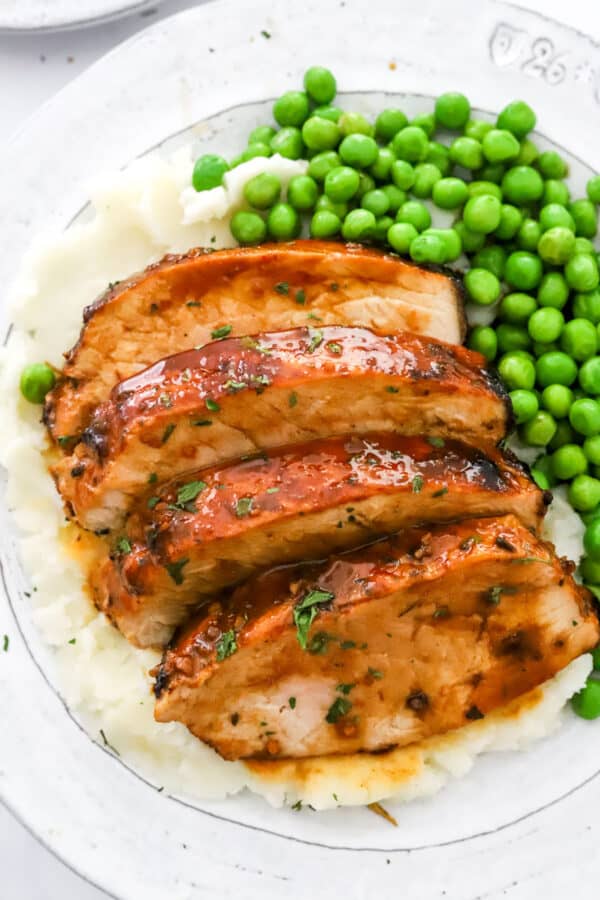 Close up shot of sliced pork loin with glaze on it on a plate with mashed potatoes and peas.