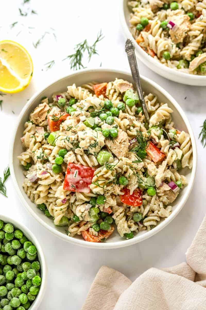 Bowl filled with tuna pasta salad with peas and peppers in it with a spoon in the bowl with another bowl of the salad behind it. 