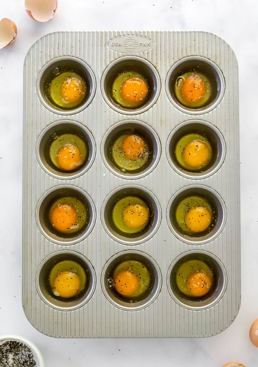 Uncooked cracked eggs in a grey muffin pan with egg shells in front of an behind it.