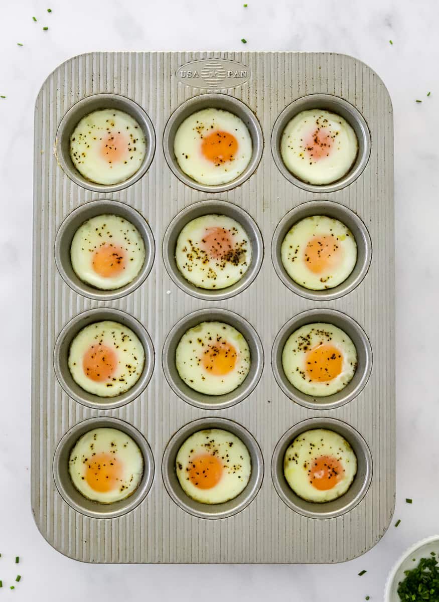Baled whole eggs in a silver muffin pan with a bowl of chopped chives in front of it.