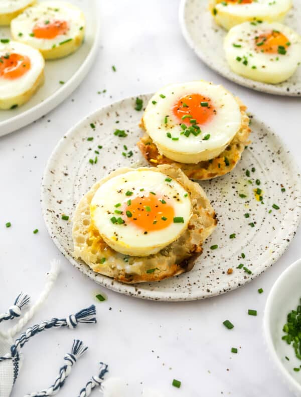 Round white and brown plate with a toasted, sliced English muffin on it with a whole cooked eggs on each half of the muffin with more baked eggs behind it.