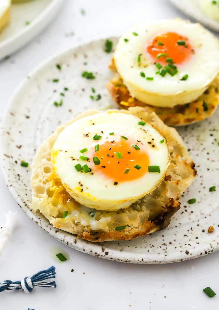 Round plate with oven baked egg on a round, toasted muffin with chopped herbs sprinkled on top of the egg and another egg on a muffin behind it.