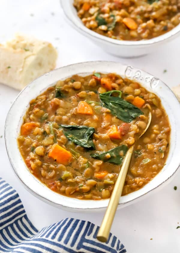 Bowl of lentil vegetable soup with a gold spoon in the bowl with another bowl of lentil soup and baguette behind it.