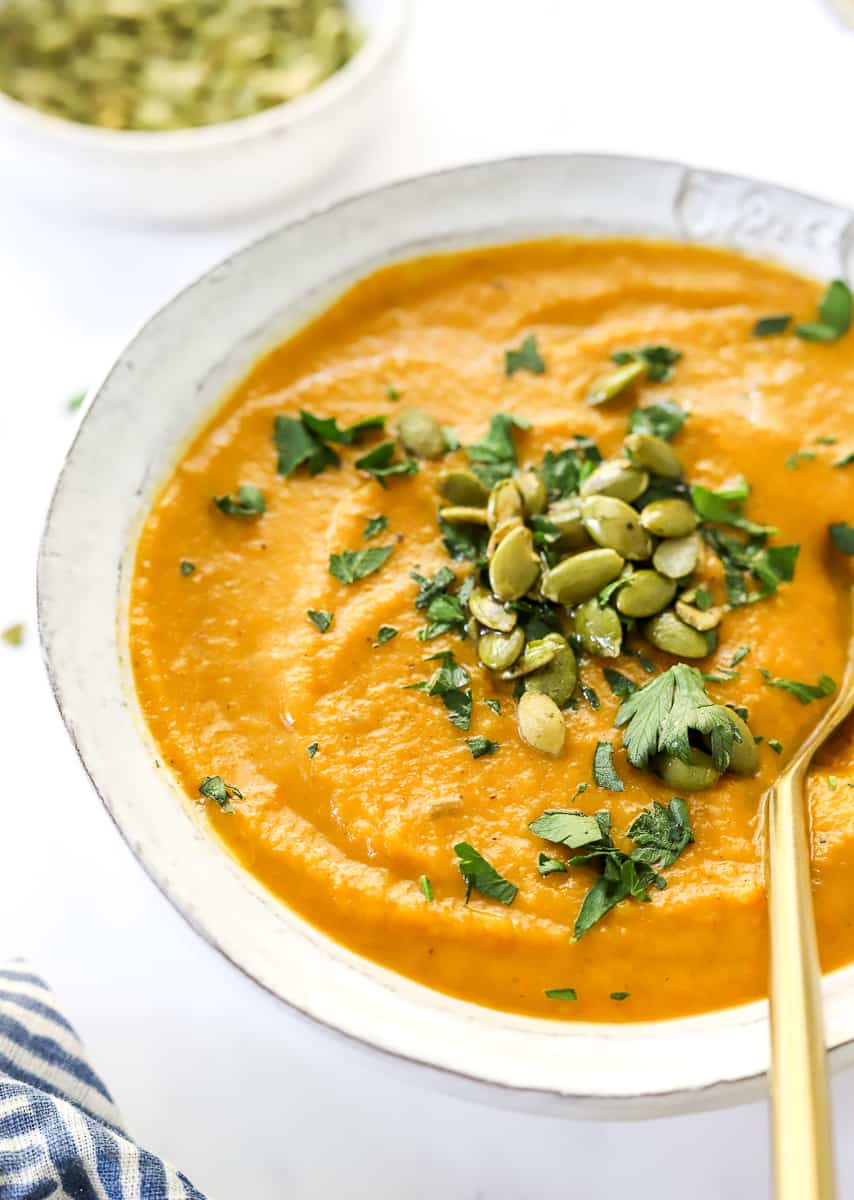 Creamy vegan butternut squash soup in a bowl with pumpkin seeds and parsley on it with a gold spoon in the bowl. 