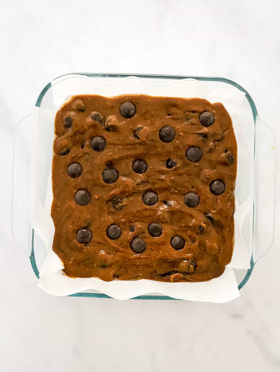 Uncooked brownie batter with chocolate chips on top in a square, glass baking pan lined with parchment paper.