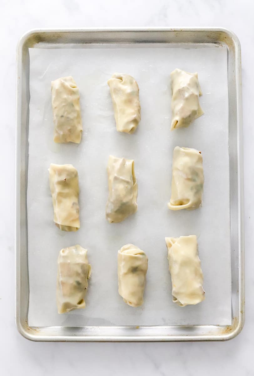 Uncooked rolled egg rolls on a lined baking sheet.