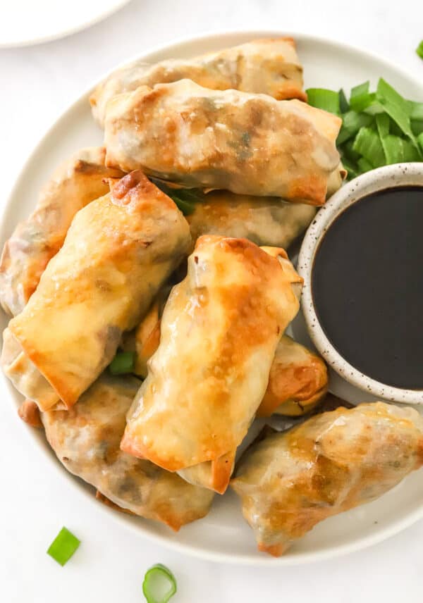 Round plate with a pile of egg rolls on it with a bowl of soy sauce an some chopped green onions next to it.