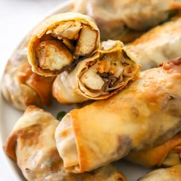 Egg rolls filled with tofu and veggies in a pile on a plate with two sliced open on top.