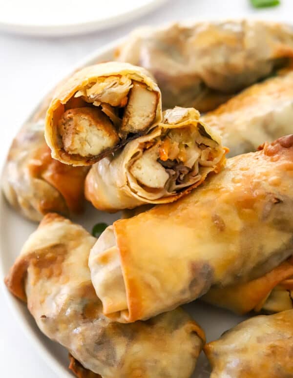 Egg rolls filled with tofu and veggies in a pile on a plate with two sliced open on top.