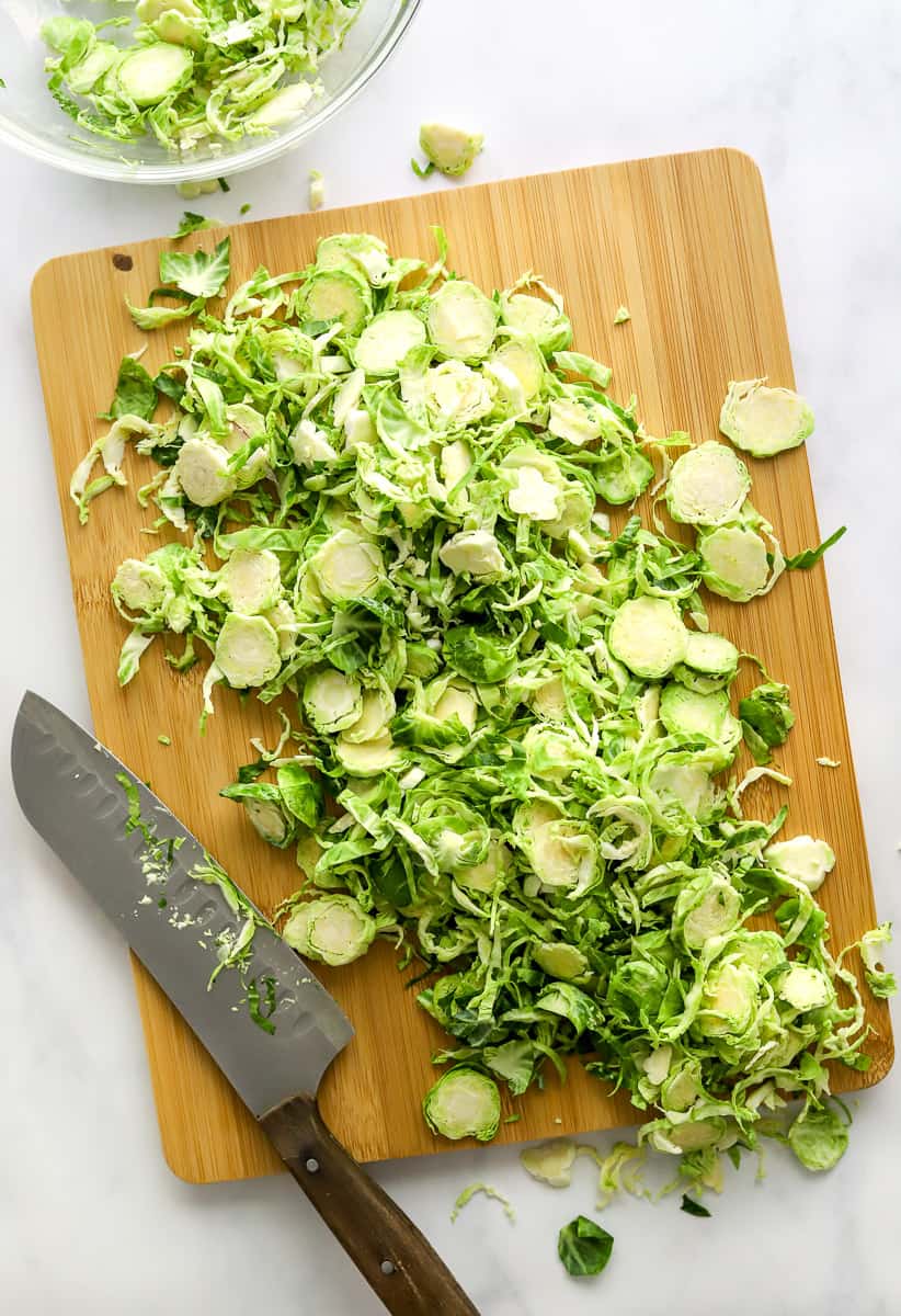 Thinly sliced Brussel Sprouts on a bamboo cutting board with a knife on the cutting board and a bowl of more sliced Brussel Sprouts behind it.