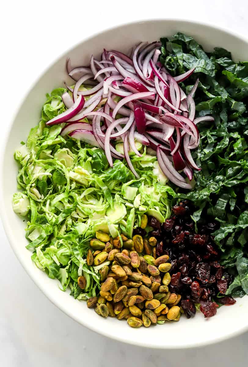 Shredded Brussel Sprouts, sliced kale, sliced red onion, dried cranberries and pistachios in a while salad bowl.