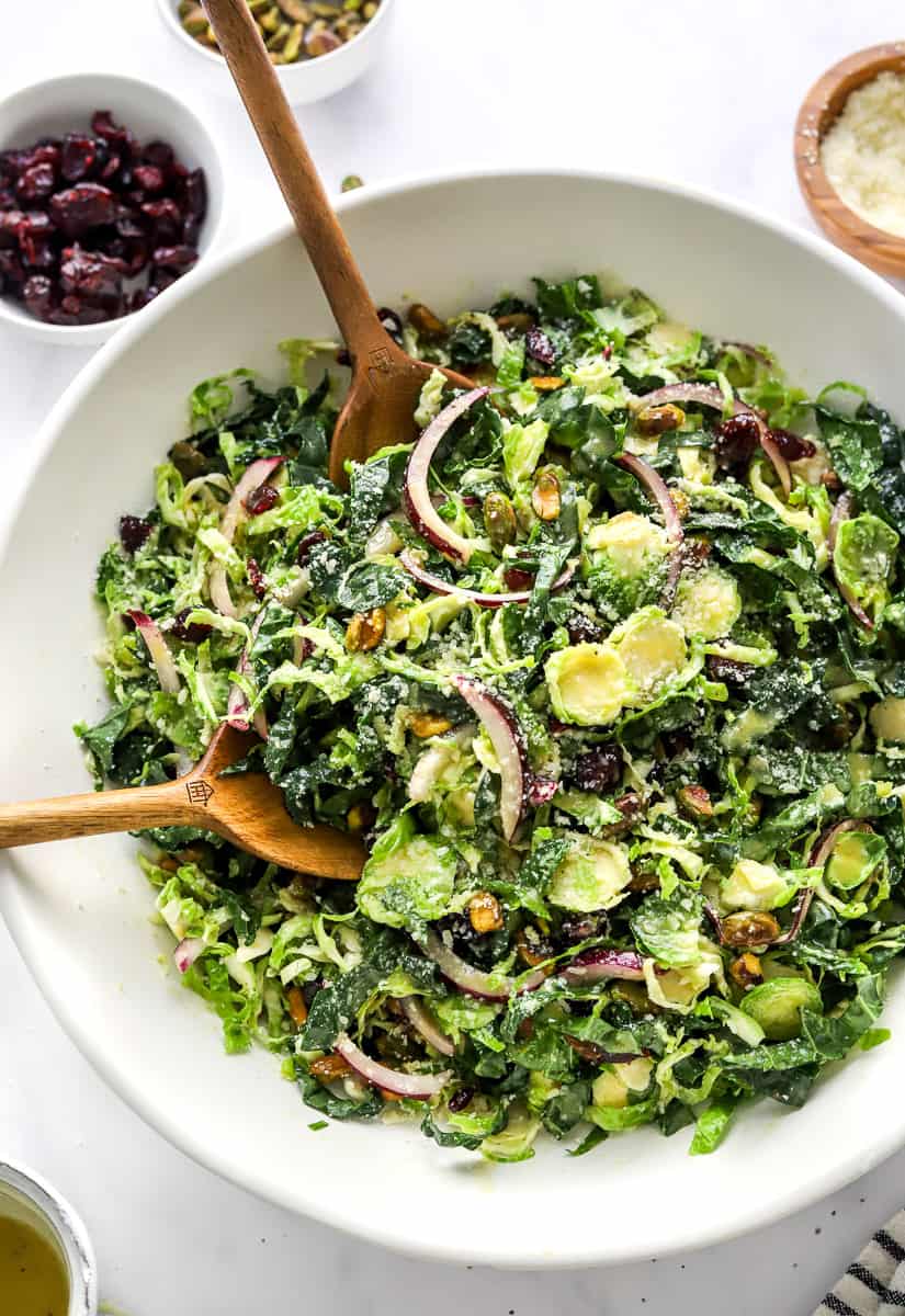 Large white bowl full of kale salad with wooden serve-ware in the bowl with a bowl of dried cranberries and a bowl of pistachios and a Bowl of parmesan cheese behind it.