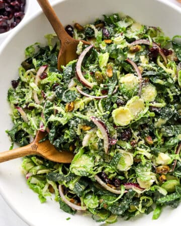 Shredded brussel sprout, kale, onion and nuts in a serving bowl with salad spoons in the bowl with a bowl of dried cranberries behind it.