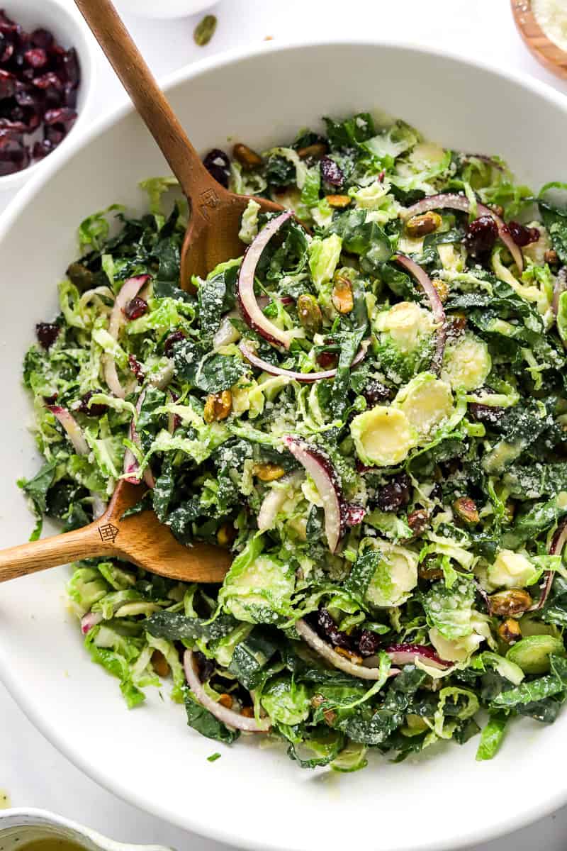 Shredded brussel sprout, kale, onion and nuts in a serving bowl with salad spoons in the bowl with a bowl of dried cranberries behind it.