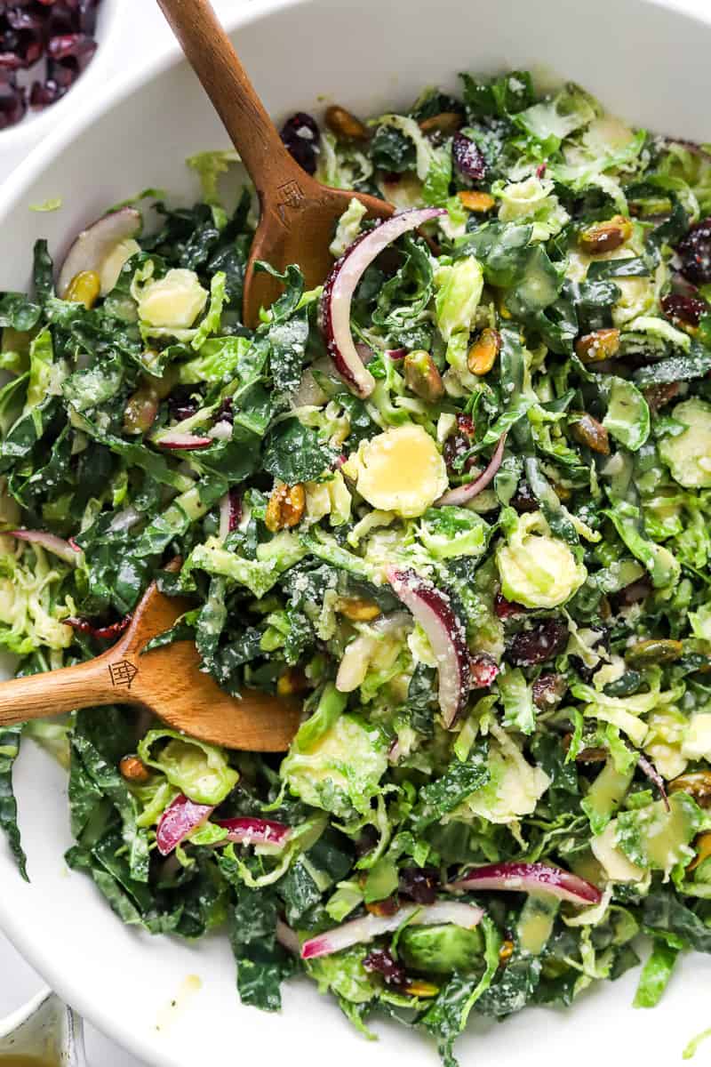 Kale and Brussel Sprouts mixed with red onion and nuts in a white salad bowl with brown serving spoons in the bowl with the salad.